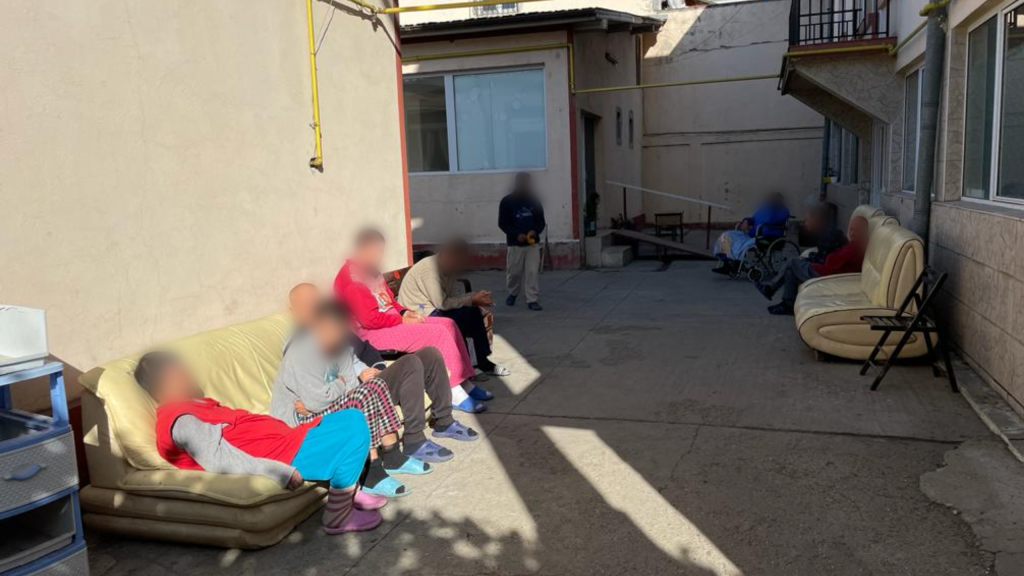 Residents in the front yard. Visibly distressed, in tracksuits and flipflops. The yard is very dark and untidy, cemented and unclean.